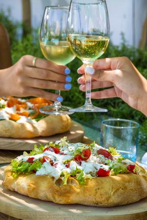 Pizza gourmet con verdure e brindisi con vino bianco all'aperto.